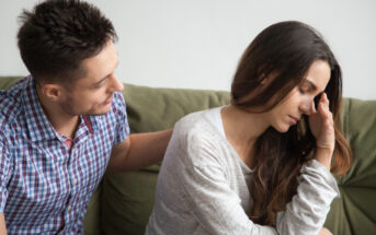 A man with short dark hair sits on a couch next to a woman with long dark hair. The woman looks distressed, with her hand covering part of her face. The man appears to be comforting her by placing a hand on her shoulder. They are indoors, in a well-lit room.