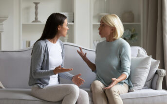 Two women, one younger with dark hair and one older with blonde hair, are sitting on a beige couch in a living room. They are engaged in a heated discussion, with both gesturing emphatically. Light-filled shelves and decorations are visible in the background.