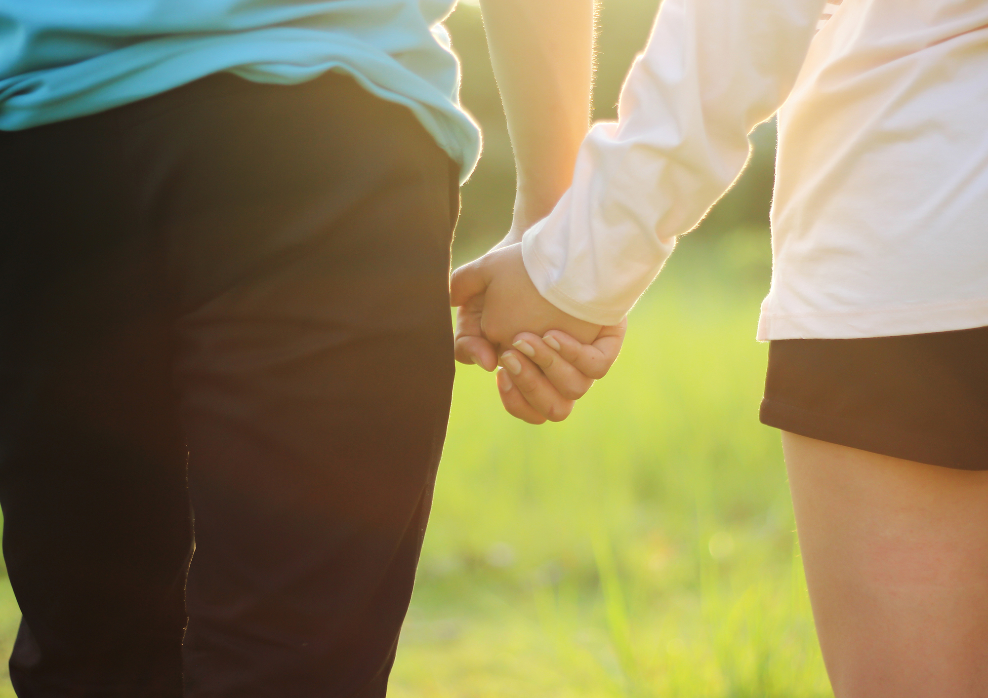 A close-up of two people holding hands while standing outdoors. The person on the left is wearing dark pants and a green shirt, and the person on the right is wearing black shorts and a white top. The background is a grassy field lit with warm sunlight.