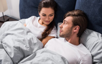 A man and woman lie in bed together under grey blankets, both wearing white shirts. They face each other, the woman smiling softly while the man gazes at her. The background is dark blue with a nightstand visible. The atmosphere appears calm and intimate.