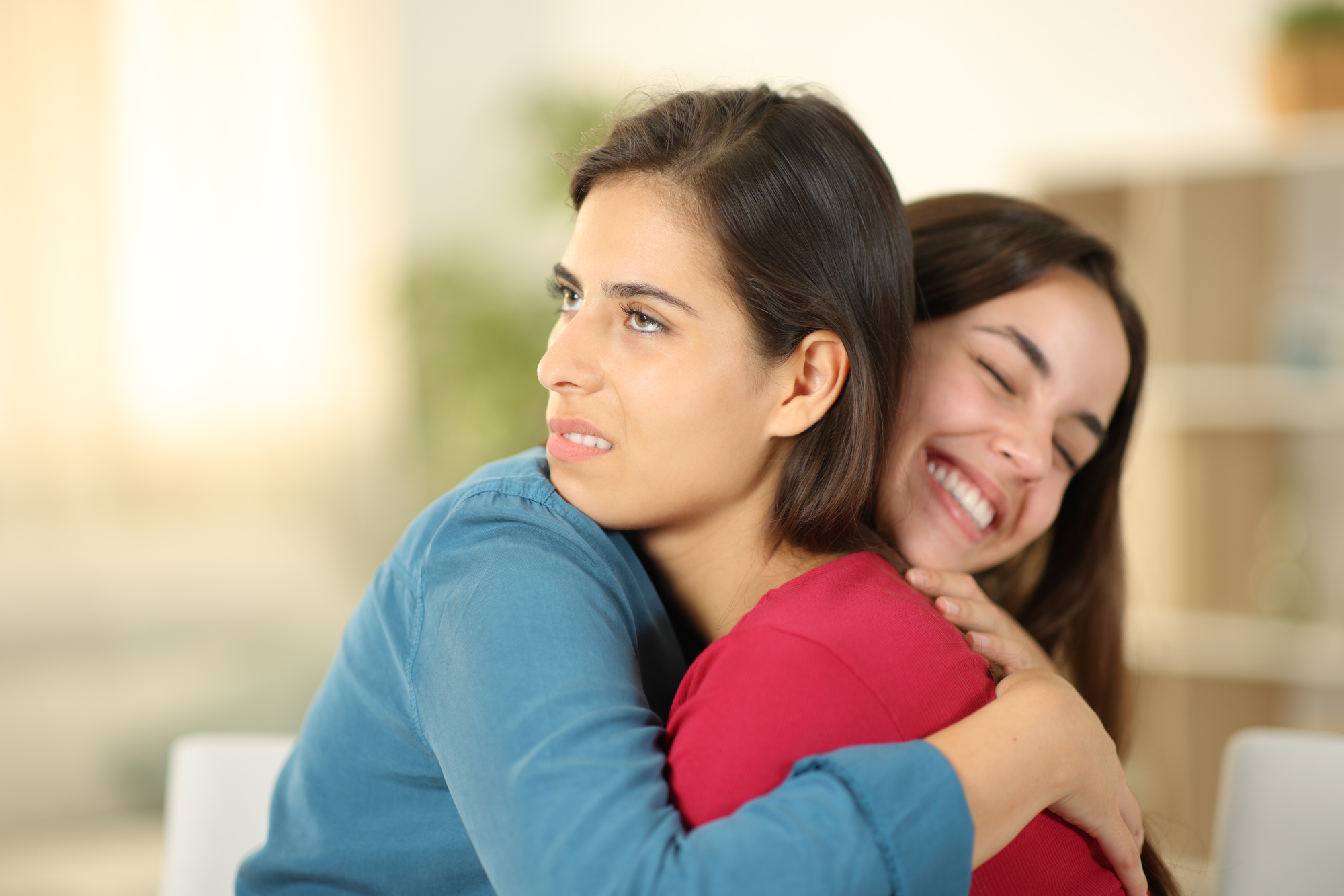 Two women are hugging indoors. The woman on the right, wearing a red shirt, has a big smile and appears happy. The woman on the left, wearing a blue shirt, looks uncomfortable and is grimacing. The background is out of focus, showing a light and airy room.