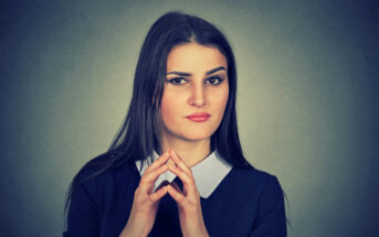 A woman with long dark hair and a serious expression is looking directly at the camera. She is wearing a dark top with a white collar and has her hands together in front of her face, fingers intertwined. The background is plain and dark.