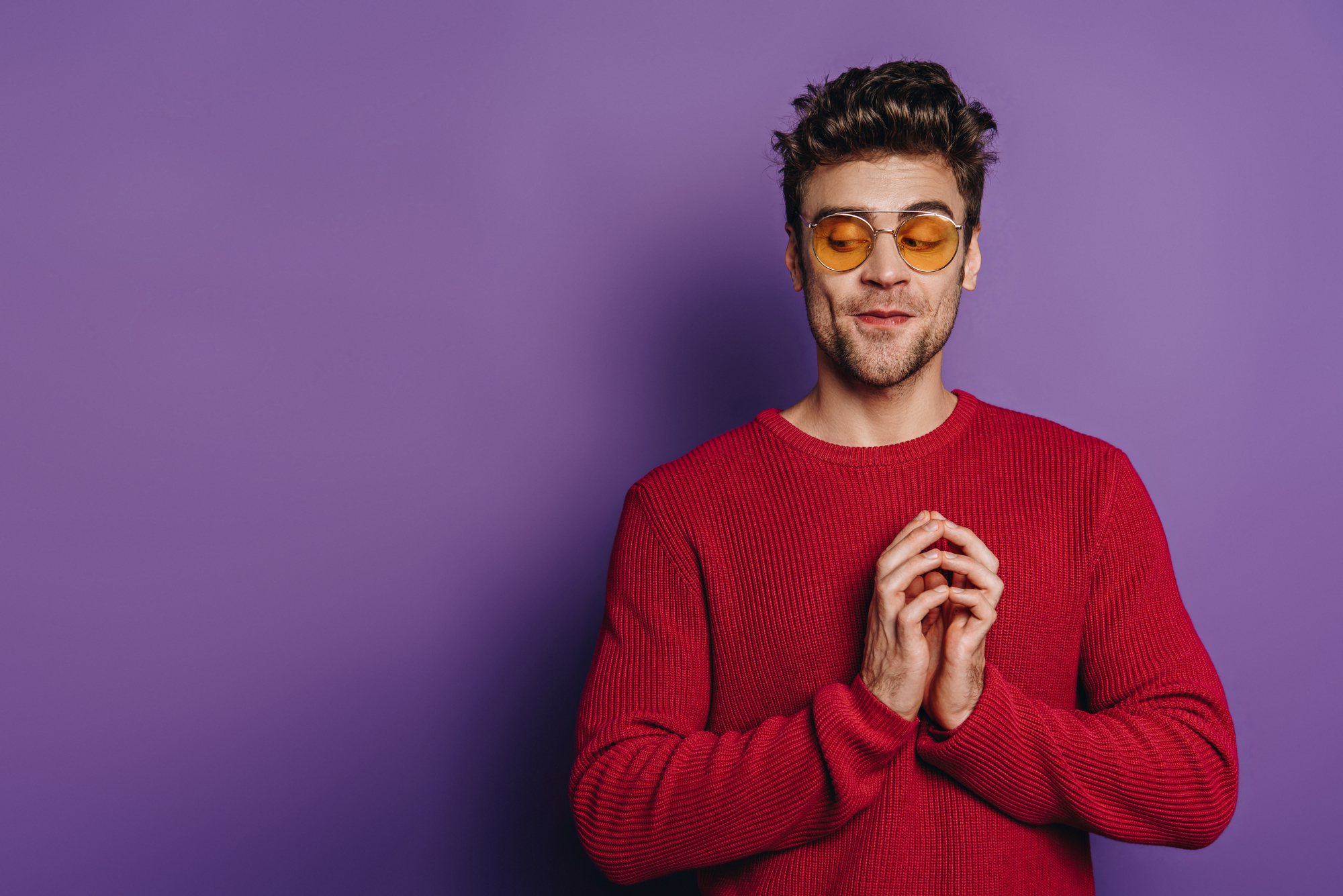 A man with short dark hair and a beard wearing orange-tinted sunglasses and a red sweater stands against a purple background. He has a slight smile and his hands are held together near his chest.