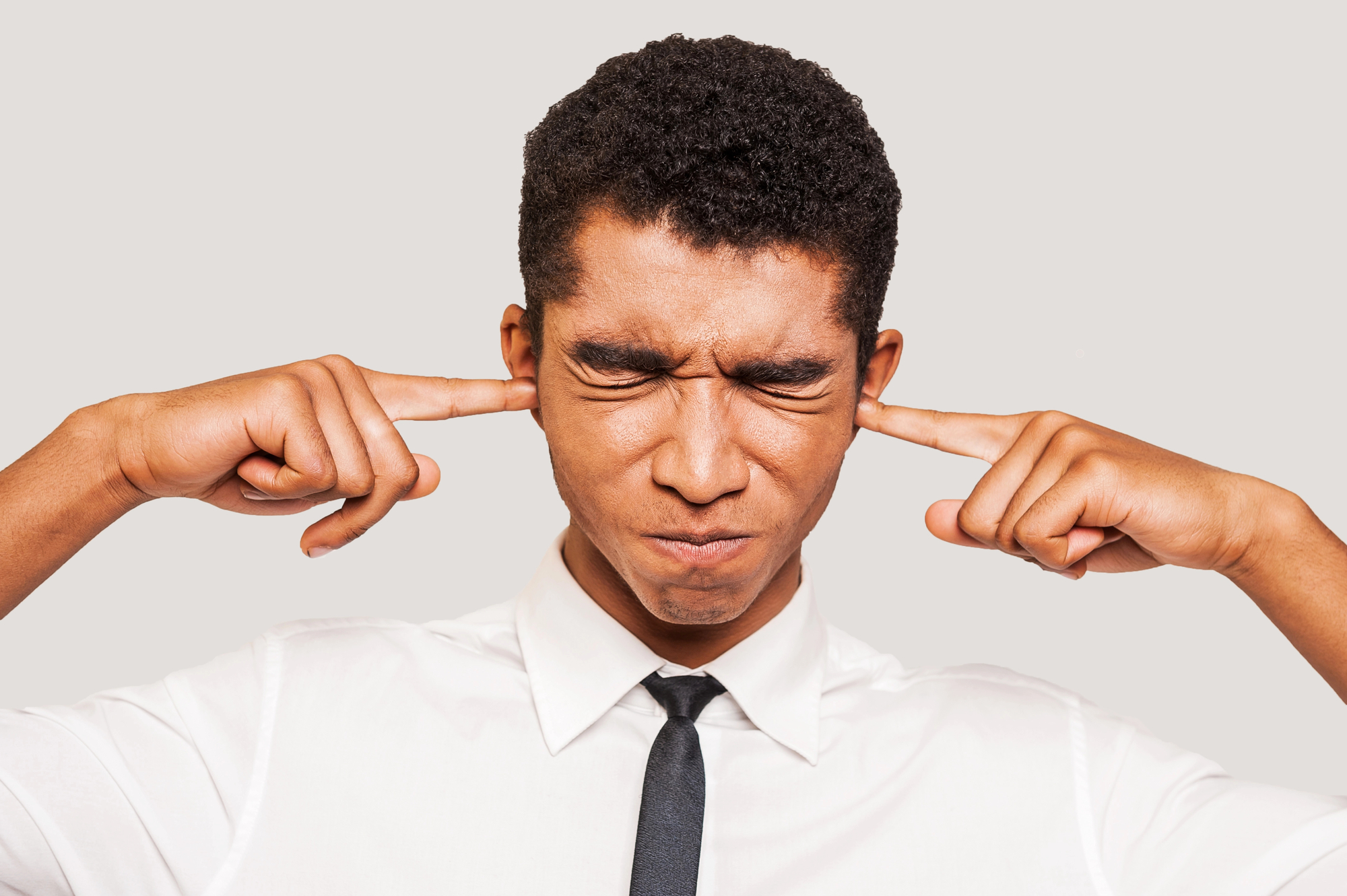 A man in a white shirt and black tie is closing his eyes and plugging his ears with his fingers. His facial expression appears strained or uncomfortable, and he is standing against a plain, light-colored background.