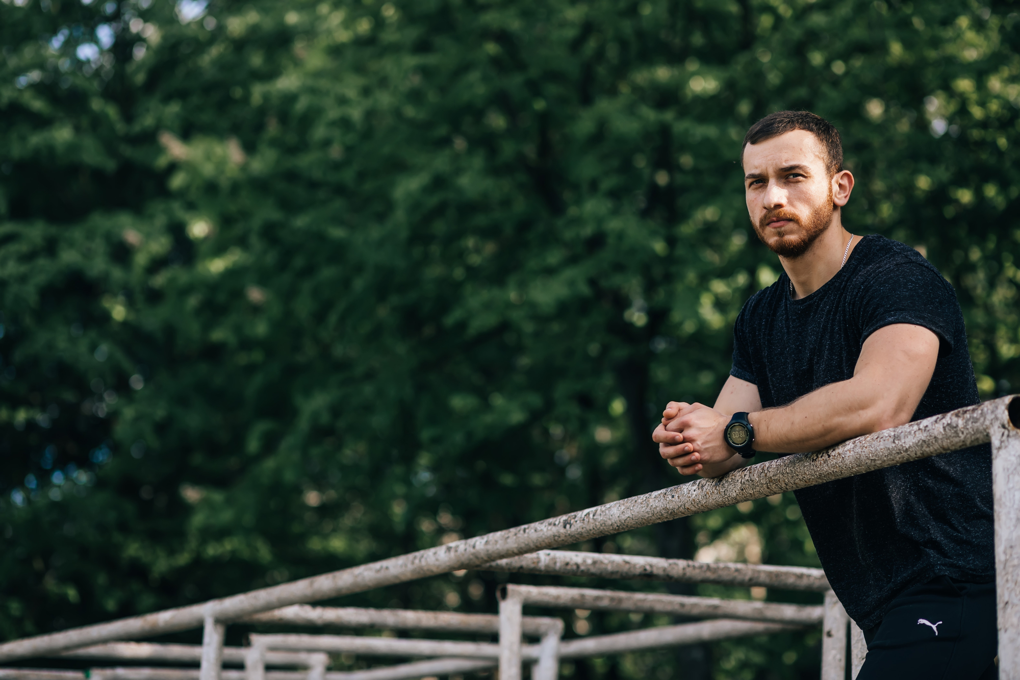 A man in athletic clothing leans on an outdoor railing in a park filled with lush green trees. He looks off into the distance with a thoughtful expression. The background is blurry, emphasizing the serene, natural setting.