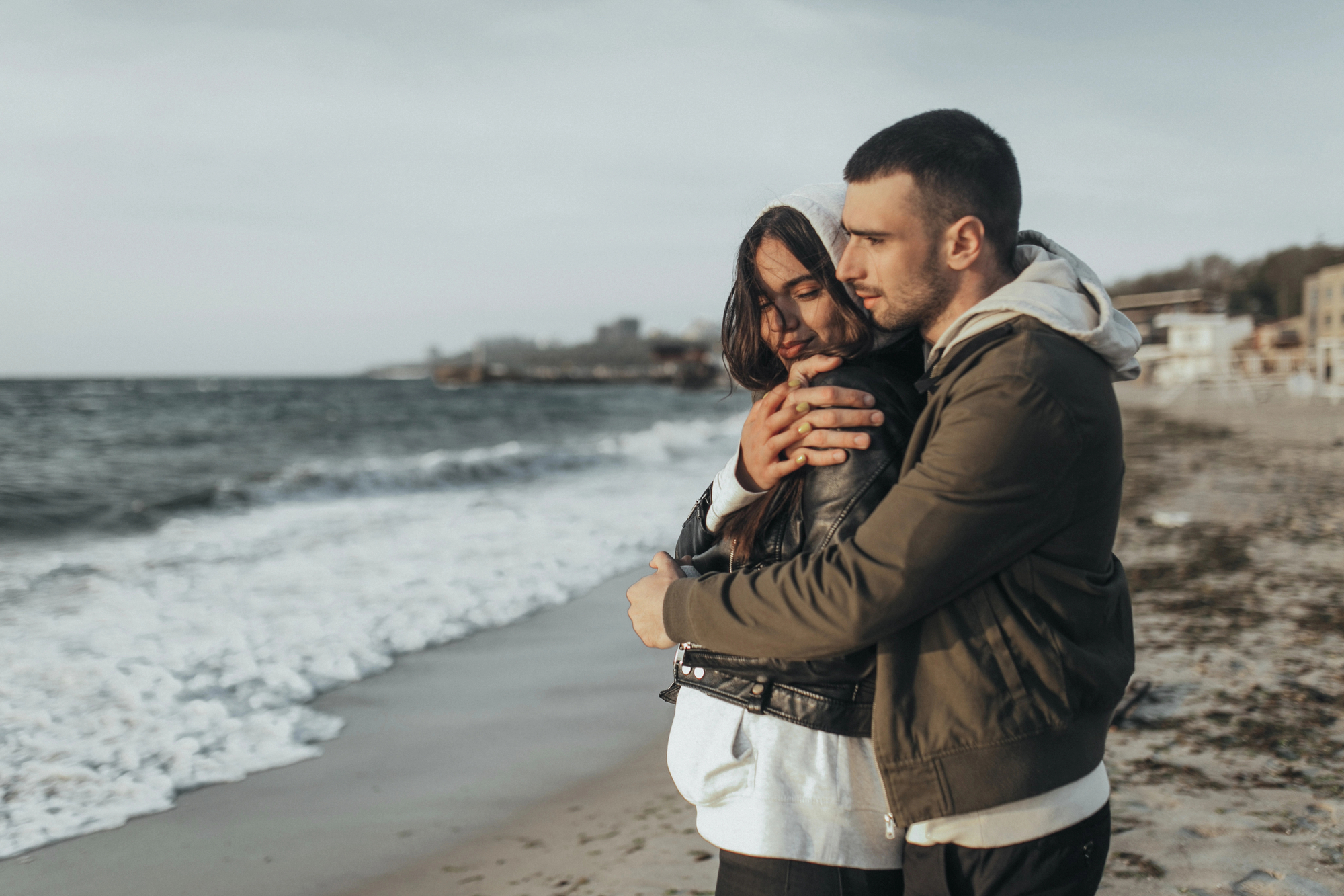 A couple embraces on a beach with waves in the background. The person on the right, wearing a green jacket, holds the other person, who is in a black leather jacket and hoodie, from behind. They both look calm and content on the sandy shore.