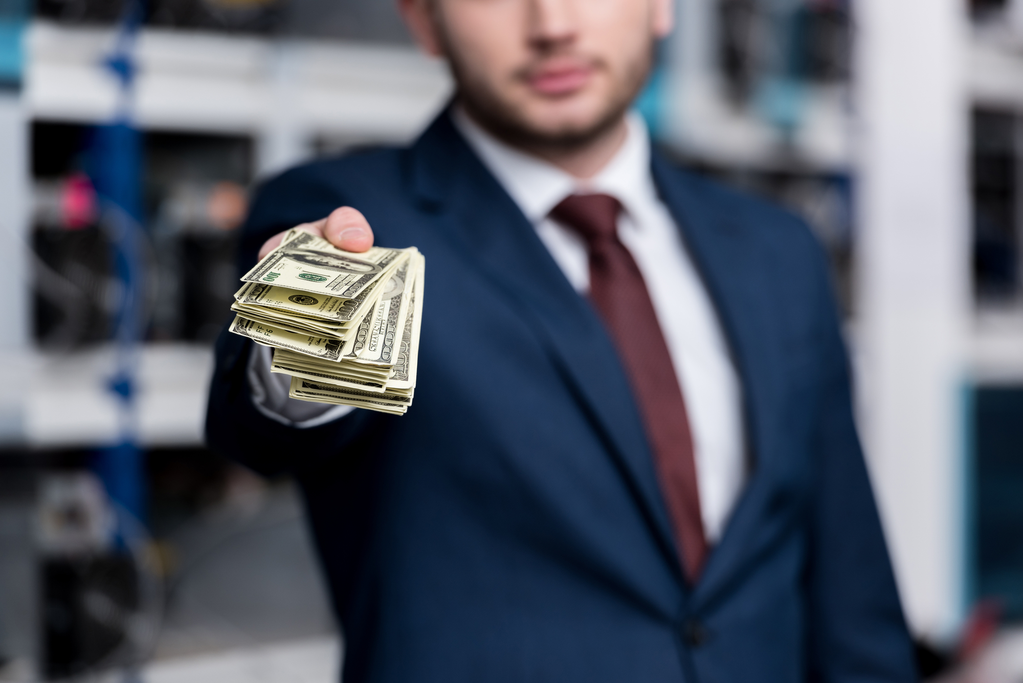 A man in a suit extending a fan of U.S. dollar bills towards the camera, with a slight blur on his face and background. The focus is on the money, emphasizing the action of offering or presenting the cash.