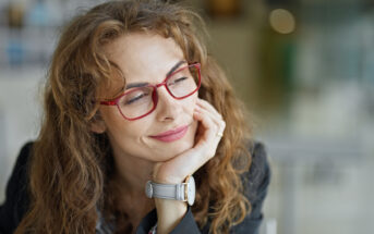 A woman with wavy brown hair wearing red glasses and a gray watch rests her chin on her hand and gazes thoughtfully to the side. She is dressed in a black blazer with a blurred background, suggesting an indoor setting.