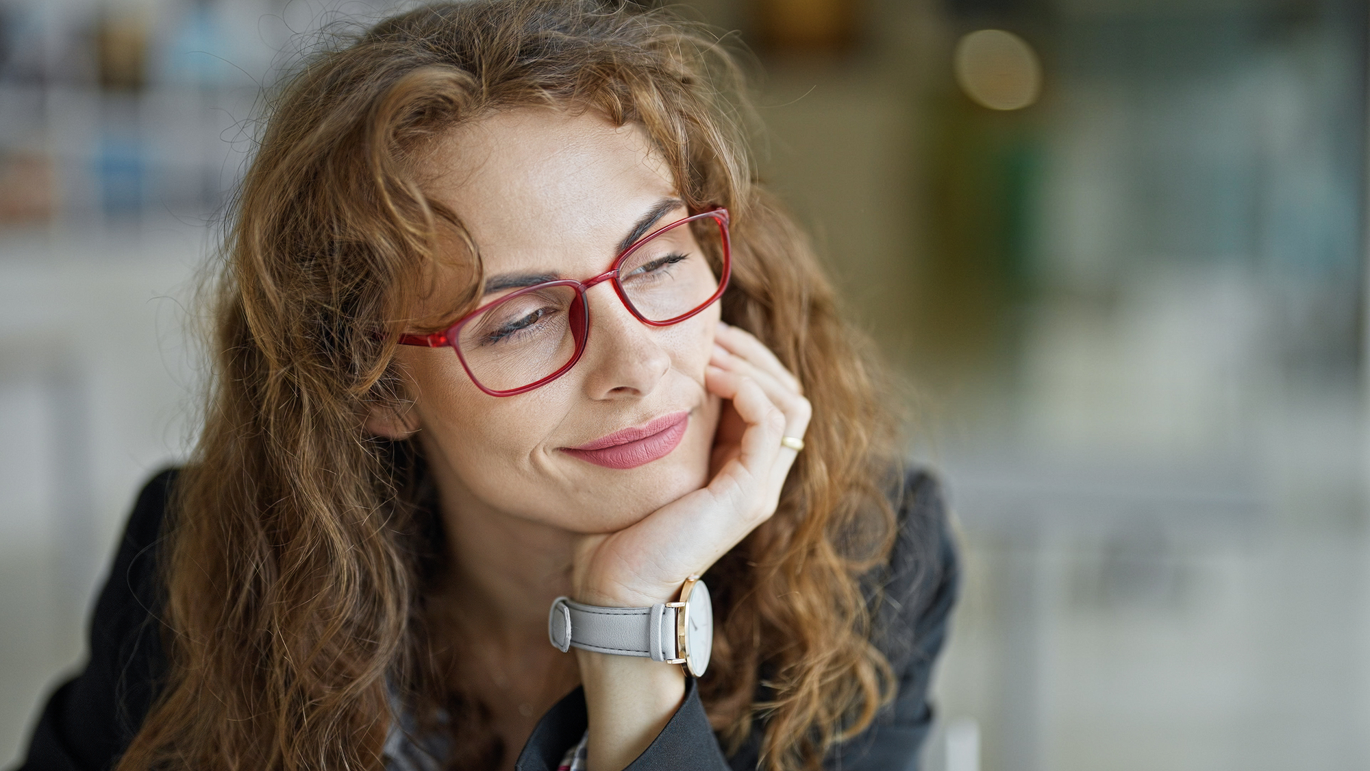 A woman with wavy brown hair wearing red glasses and a gray watch rests her chin on her hand and gazes thoughtfully to the side. She is dressed in a black blazer with a blurred background, suggesting an indoor setting.