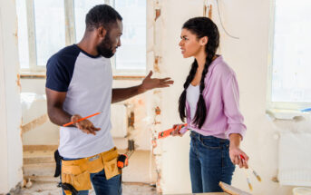 A man and woman are arguing in a partially renovated room. The man has a pencil in hand and is wearing a tool belt, while the woman, holding a paintbrush and measuring tape, looks frustrated. Both are standing near a table with renovation tools.