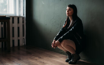 A woman with long dark hair sits on her heels against a dark green wall in a dimly lit room with wooden floors. She is wearing a black dress and jacket, with her hands resting on her knees, looking contemplative. A window with diffused light is in the background.