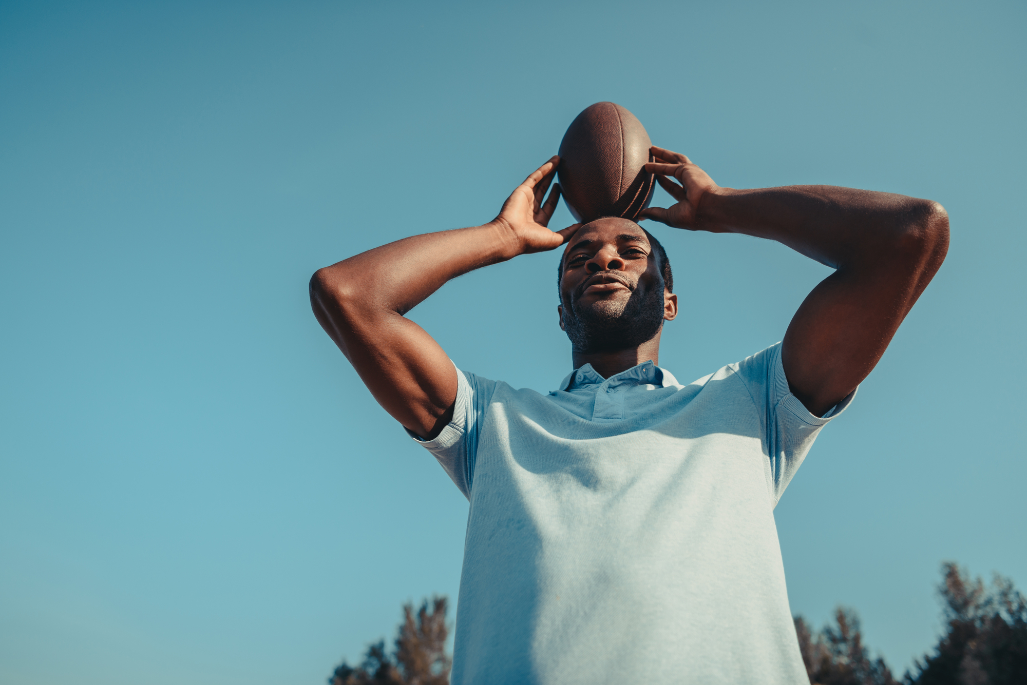 A man in a light blue shirt holds an American football above his head with both hands. He stands outdoors against a clear blue sky, with trees faintly visible in the background. His expression is calm, and the photo is taken from a low angle.