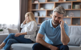 A man with a serious expression sits on a couch, leaning his head on his hand. In the background, a woman is sitting on the same couch, turned away, also appearing upset. A bookshelf with various items is in the background. The scene suggests tension or conflict.