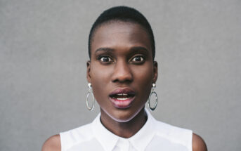 A person with a closely shaved hairstyle, wearing hoop earrings and a sleeveless white top, looks directly at the camera with a surprised or shocked expression. The background is plain and gray.