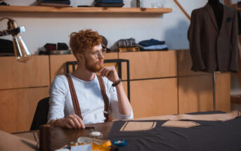 A person with curly red hair and glasses sits at a table in a tailor shop, surrounded by sewing materials and pattern pieces. They are wearing a white shirt and suspenders, gazing thoughtfully to the side. Clothing and fabric are visible in the background.