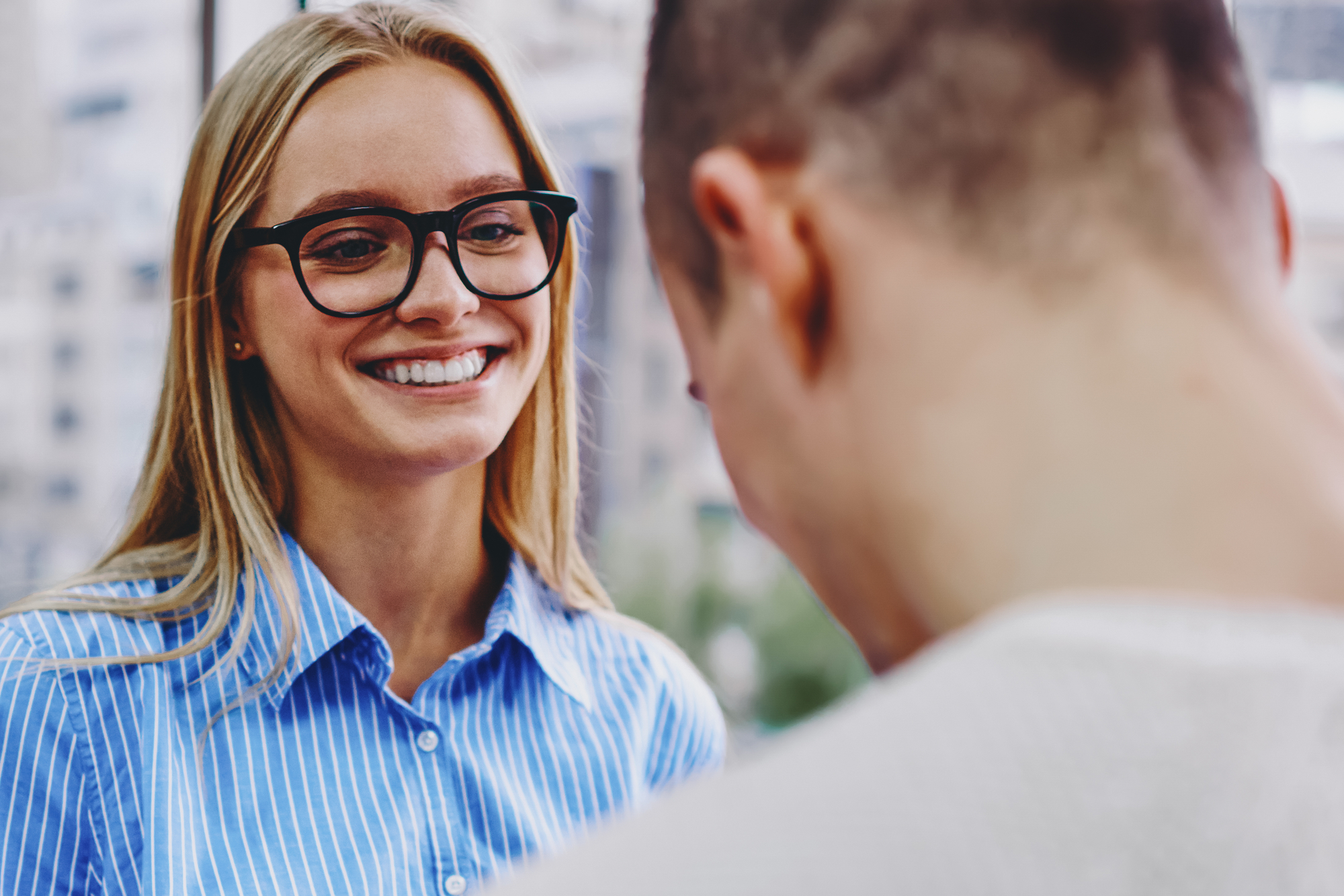 A person with long blond hair, wearing glasses and a blue shirt, is smiling while engaging in a conversation with another person who has short hair and is turned away from the camera. The background shows an urban setting with blurred buildings.