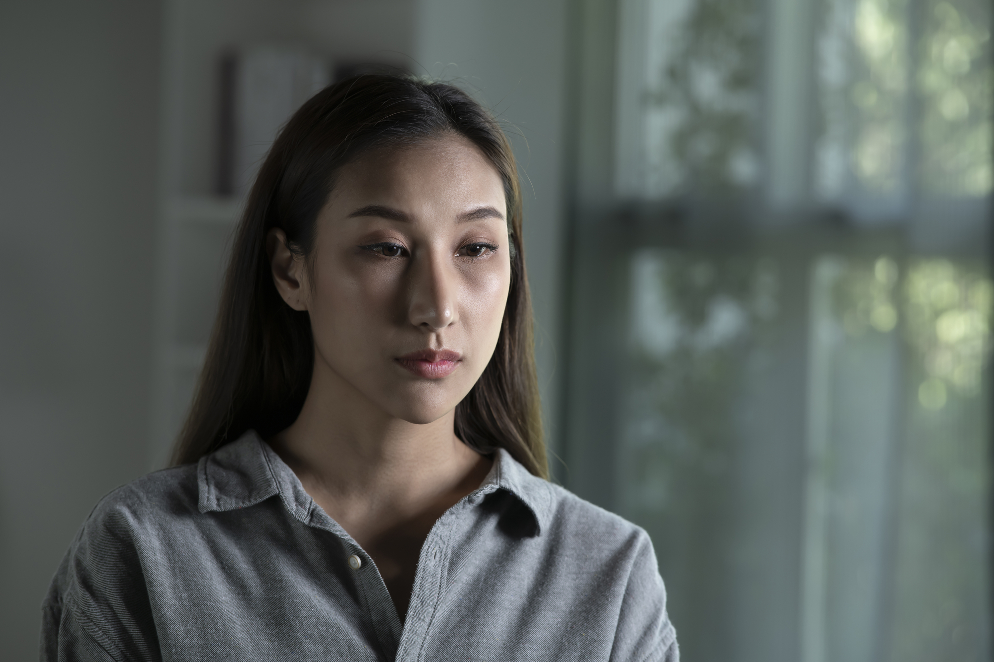 A young woman with long brown hair and a neutral expression sits indoors. She is wearing a grey button-up shirt, and the background shows a softly lit room with large windows and faint outlines of greenery outside.