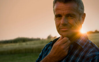 A man with short gray hair and a slight smile stands outdoors in a field at sunset. He is wearing a blue and black checkered shirt and touches his chin with his left hand. The sun is behind him, casting a warm glow. Trees and a grassy field are visible in the background.