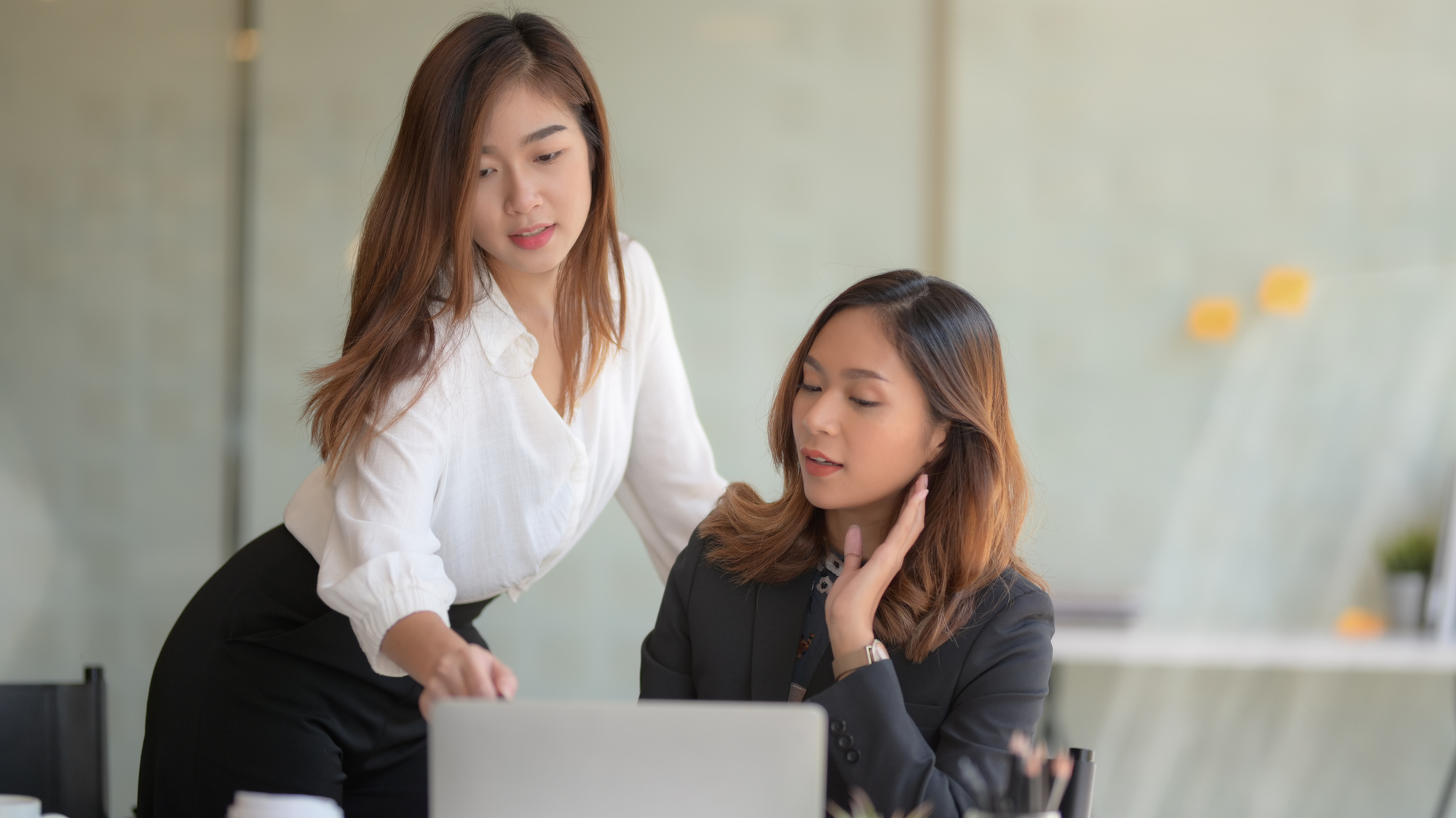 Two women are working together in an office setting. One woman is standing and pointing at a laptop screen while the other is seated, attentively looking at the screen and touching her neck. Both appear engaged and focused on the work.