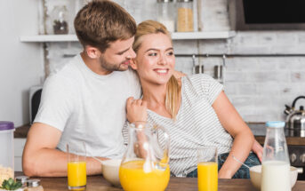 A couple sits at a kitchen counter with glasses of orange juice, a pitcher of juice, and a bottle of milk in front of them. The man is hugging the woman from the side, both smiling warmly. The kitchen background includes shelves with jars and various kitchen items.