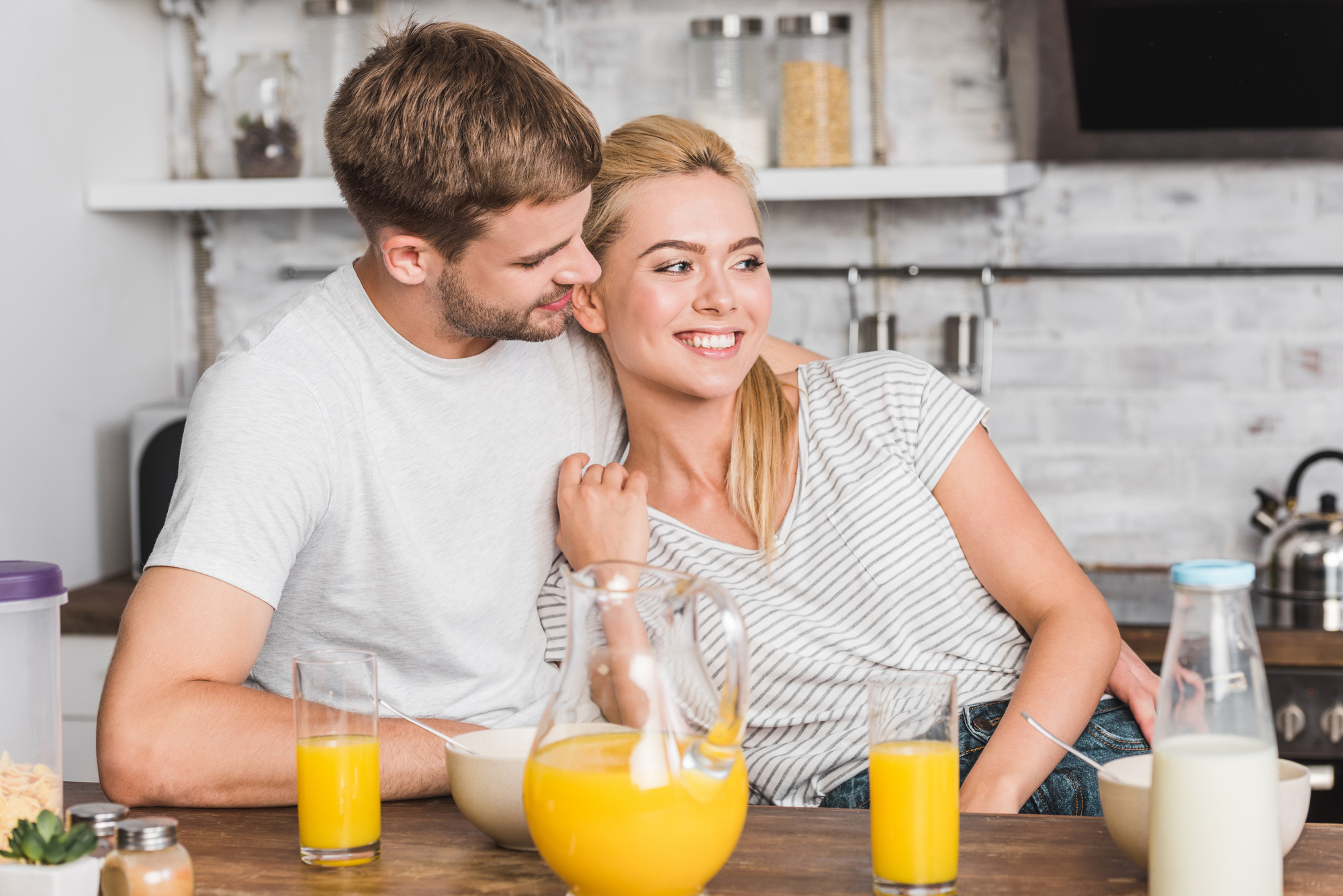 A couple sits at a kitchen counter with glasses of orange juice, a pitcher of juice, and a bottle of milk in front of them. The man is hugging the woman from the side, both smiling warmly. The kitchen background includes shelves with jars and various kitchen items.