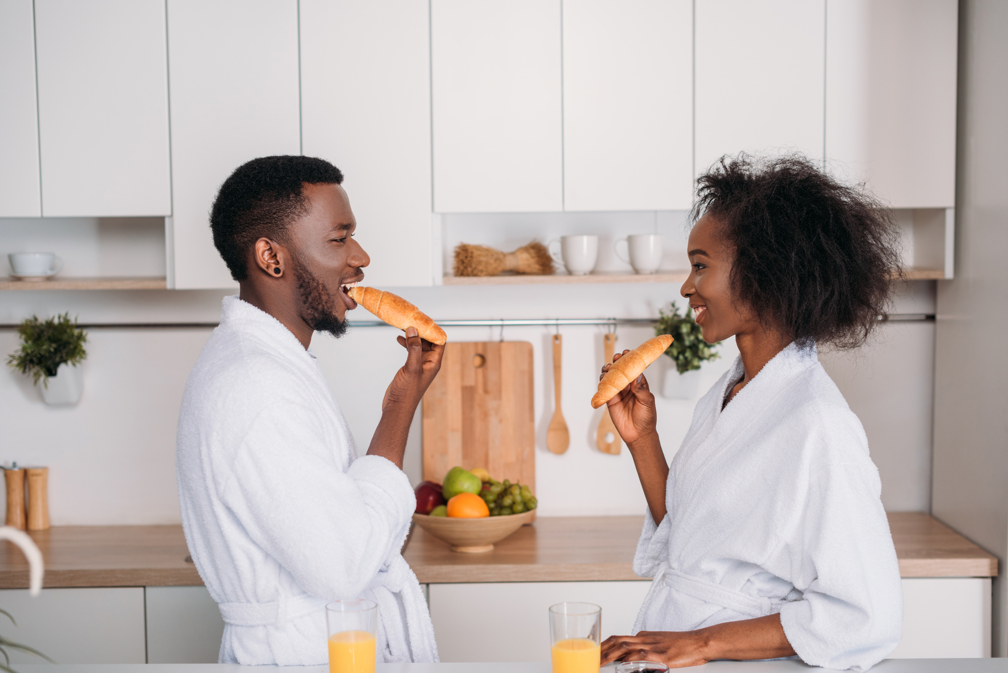 A smiling couple wearing white bathrobes stand in a kitchen, facing each other while holding croissants. They are near a countertop with a bowl of fruits and two glasses of orange juice. The white cabinets and light wood accents create a bright, modern atmosphere.