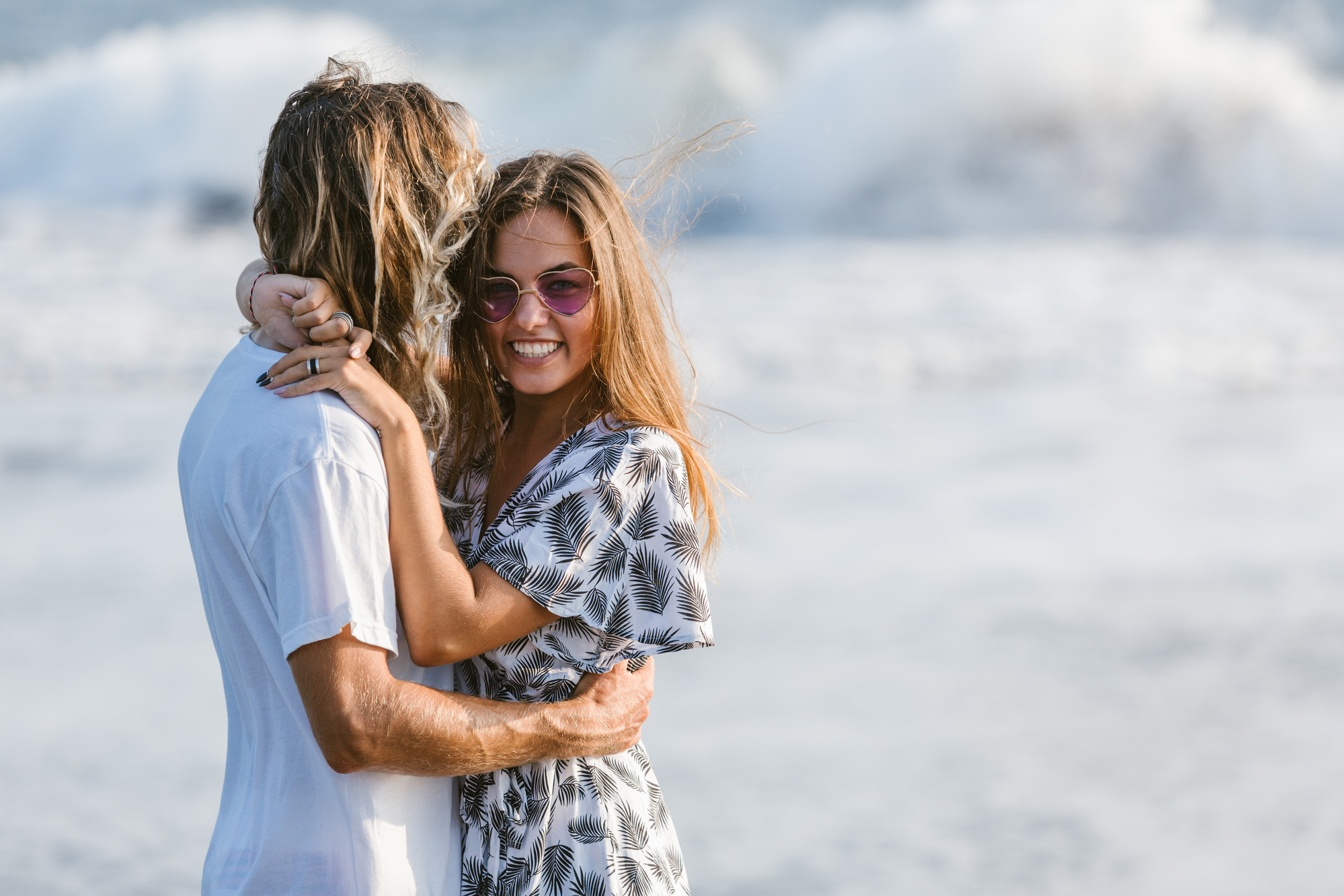 A couple embraces on a beach. The woman, wearing sunglasses and a patterned dress, smiles at the camera, while the man, with long hair and a white shirt, has his back to the camera. Waves crash on the shore in the background.