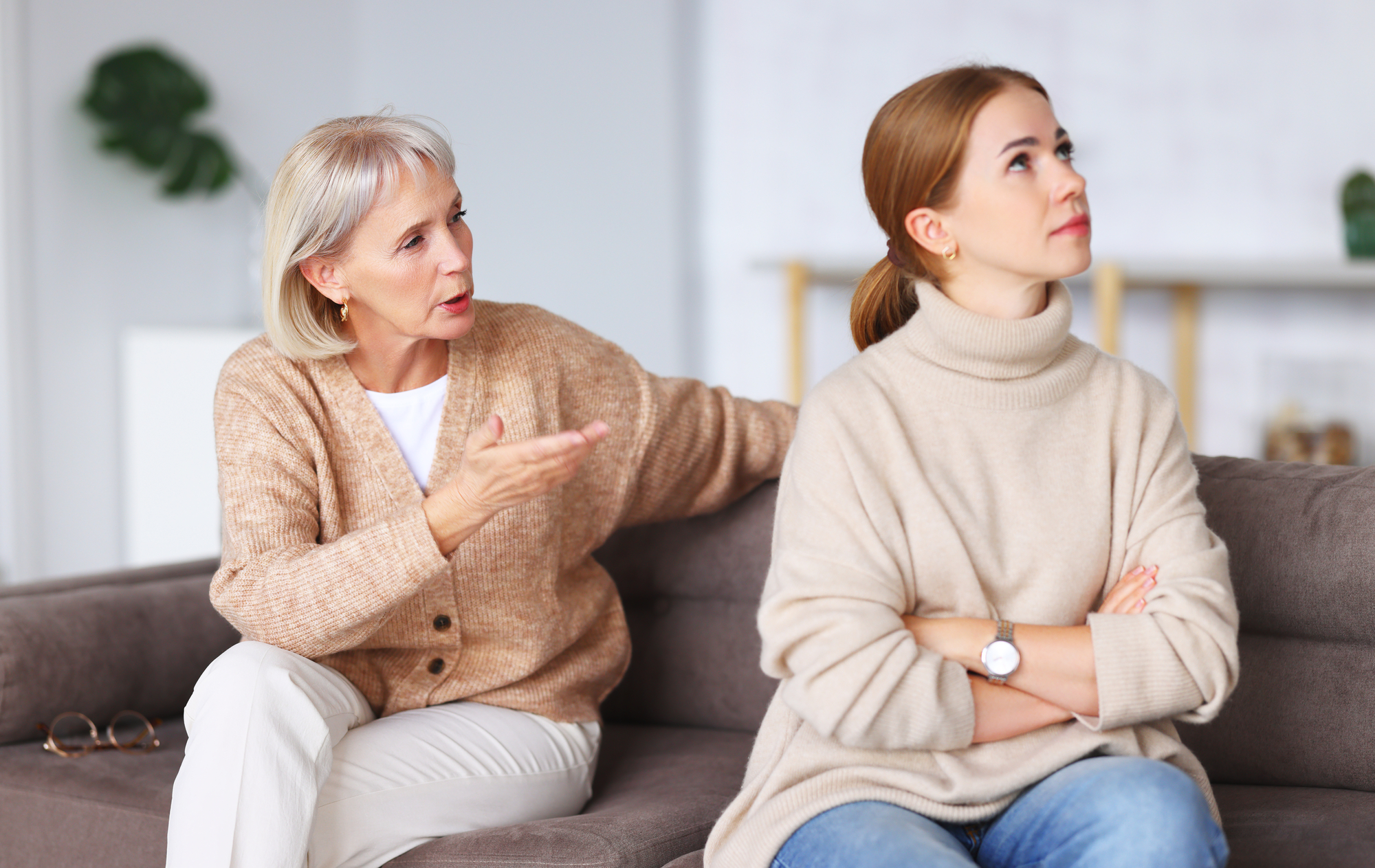 A middle-aged woman with short gray hair and a younger woman with long brown hair sit on a couch. The older woman is talking with an expressive gesture, while the younger woman looks away with her arms crossed, appearing frustrated. Both are wearing beige clothing.