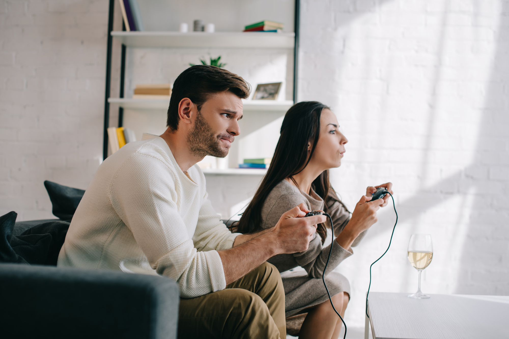 A man and woman sit on a couch intensely playing video games. Both are holding controllers, focused on the screen. There is a glass of white wine on the table in front of them. A bookcase filled with books and decorative items is visible in the background.