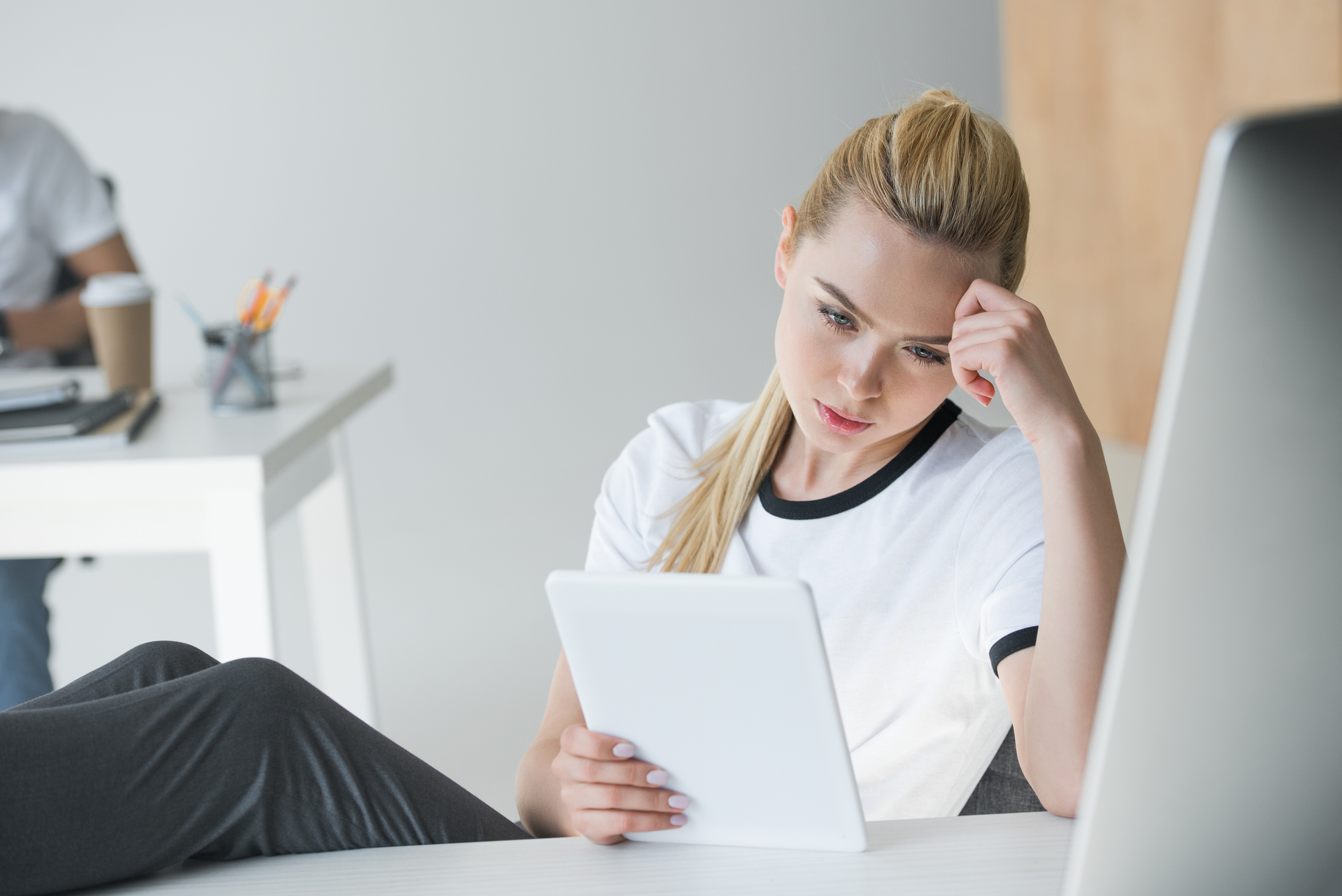 A woman with blonde hair is sitting at a desk, wearing a white t-shirt with black trim, holding a white tablet. She rests her head on one hand and appears to be deep in thought. The background features a minimalist office setting with another person in the distance.