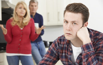 A young man in a plaid shirt, resting his head on his hand, looks frustrated in the foreground. A woman in a red cardigan and an older man stand in the background, appearing concerned, with the woman gesturing with her hands in a questioning manner.