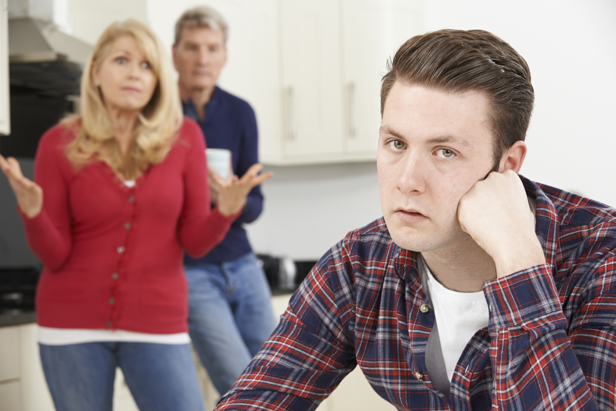 A young man in a plaid shirt, resting his head on his hand, looks frustrated in the foreground. A woman in a red cardigan and an older man stand in the background, appearing concerned, with the woman gesturing with her hands in a questioning manner.