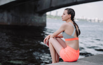 A woman in a gray sports bra and neon orange shorts sits by the water under a bridge. She has braided hair and gazes thoughtfully into the distance. The city skyline and part of the bridge's structure are visible in the background.