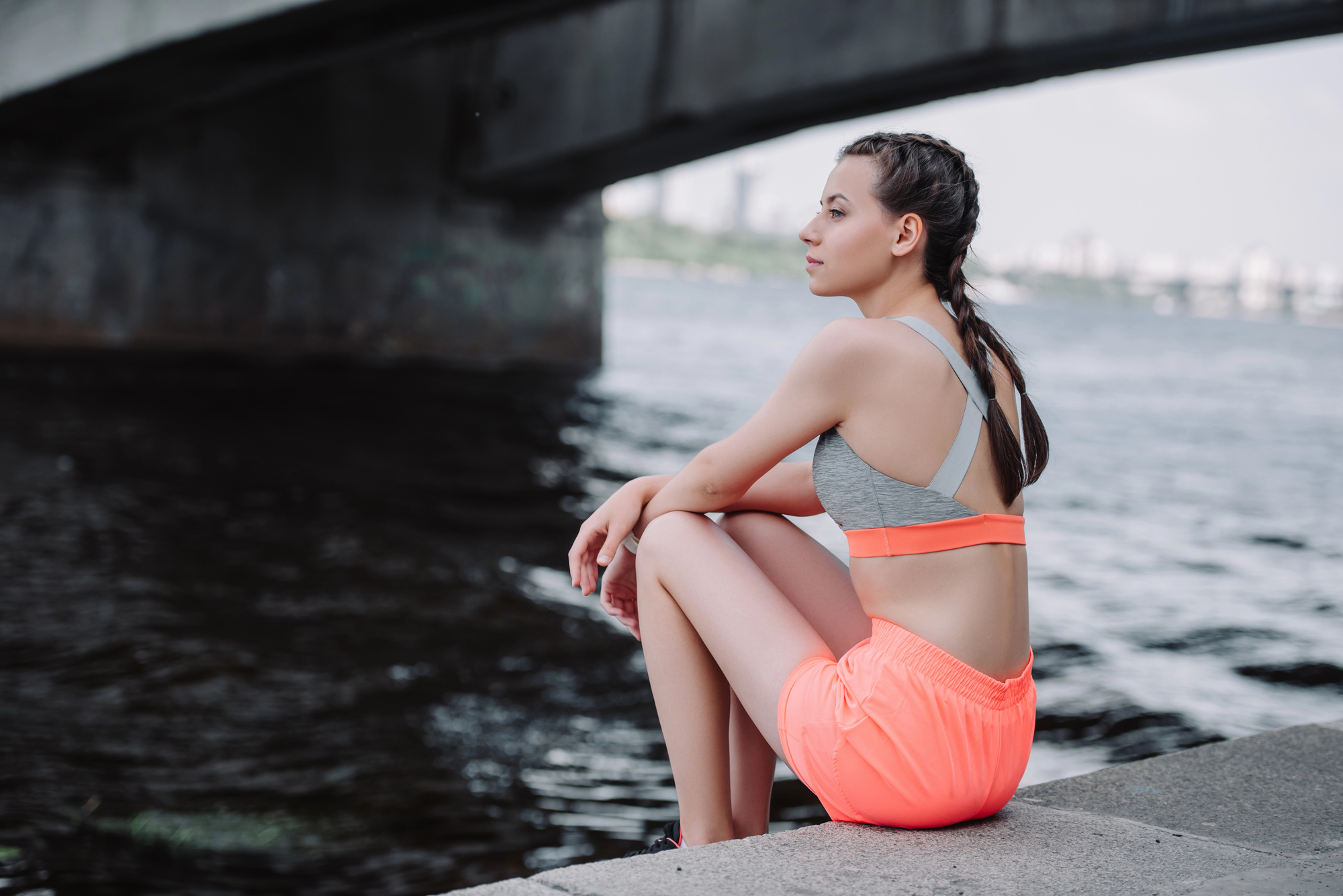 A woman in a gray sports bra and neon orange shorts sits by the water under a bridge. She has braided hair and gazes thoughtfully into the distance. The city skyline and part of the bridge's structure are visible in the background.