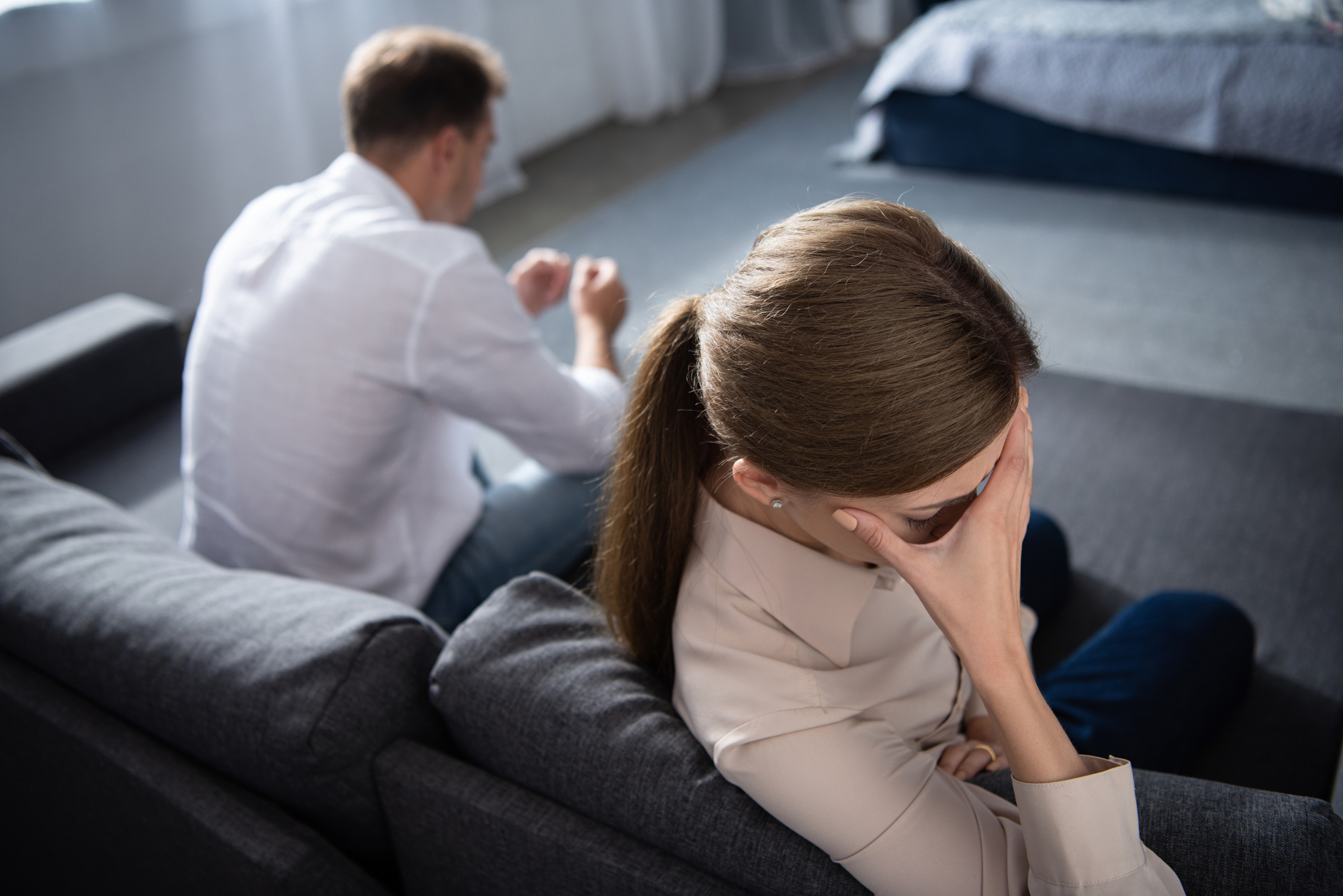 A woman sitting on a couch with her head in her hand, looking distressed, while a man sits behind her on the floor, appearing upset and looking down. They are in a room with a bed and curtains in the background.