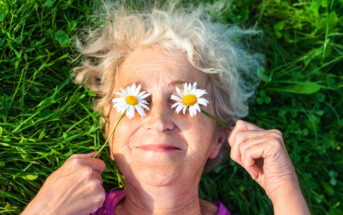 An elderly woman with gray hair lies on green grass, smiling. She holds two daisies over her eyes with both hands, creating a playful and joyful expression. The flowers contrast with her pink shirt and the lush greenery around her.