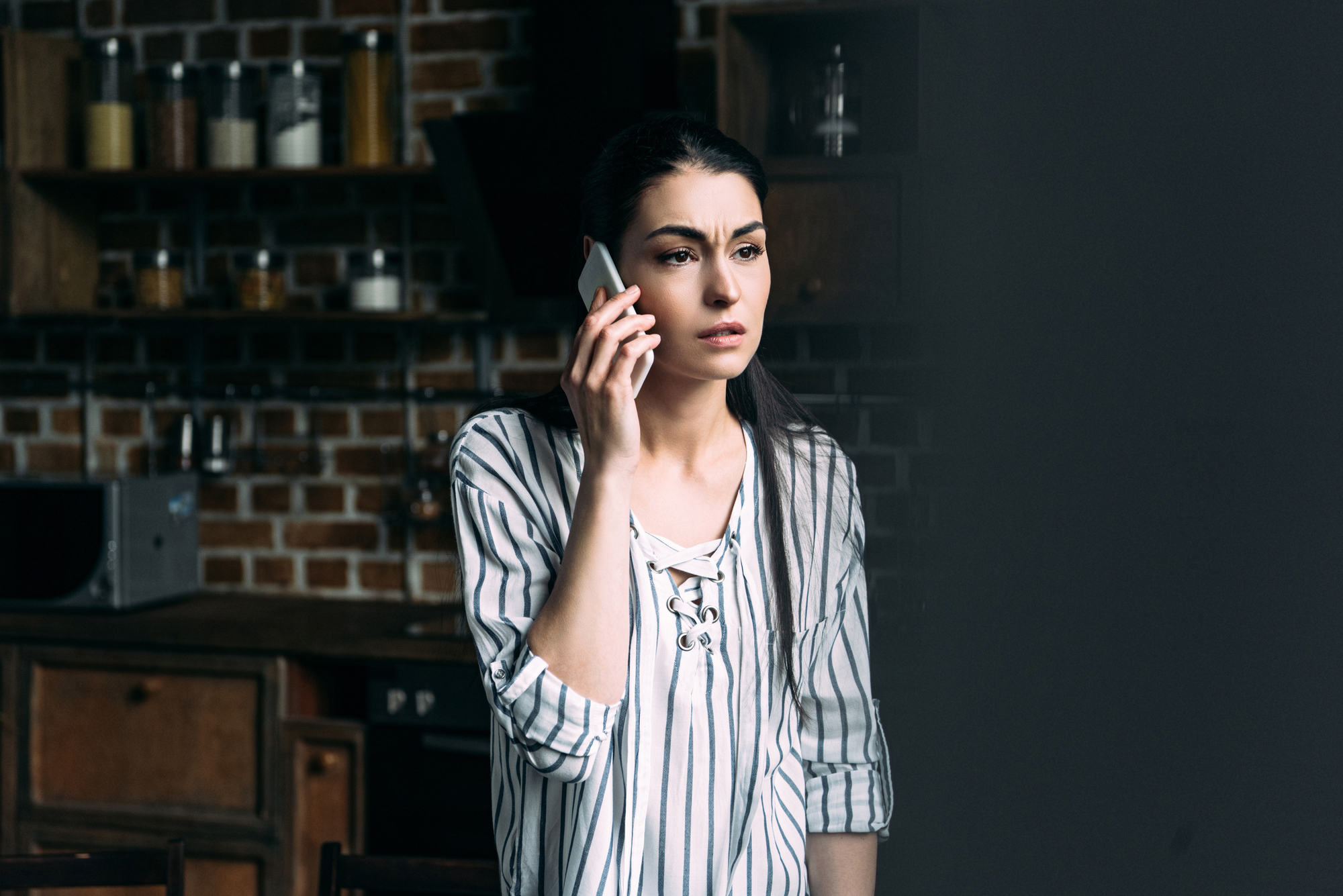 A concerned woman with long dark hair stands indoors, holding a phone to her ear. She is wearing a striped blouse and is looking off into the distance. The background is a kitchen with shelves containing jars and containers, and some wooden cabinets.