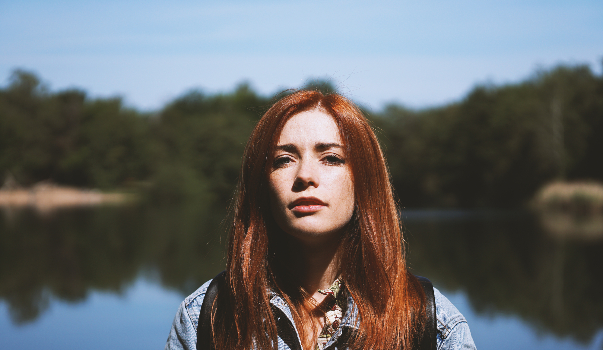 A woman with long red hair and wearing a denim jacket stands in front of a calm lake with trees in the background. She gazes directly at the camera, with a serene and thoughtful expression. The sky is clear, and the water reflects the greenery around it.