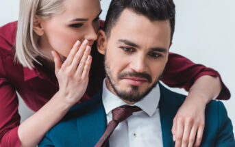 A woman with blonde hair, wearing a red shirt, leans in to whisper into the ear of a man with dark hair and a beard, who is wearing a blue suit and red tie. The man has a slight frown and is looking downwards. Both appear to be against a plain white background.