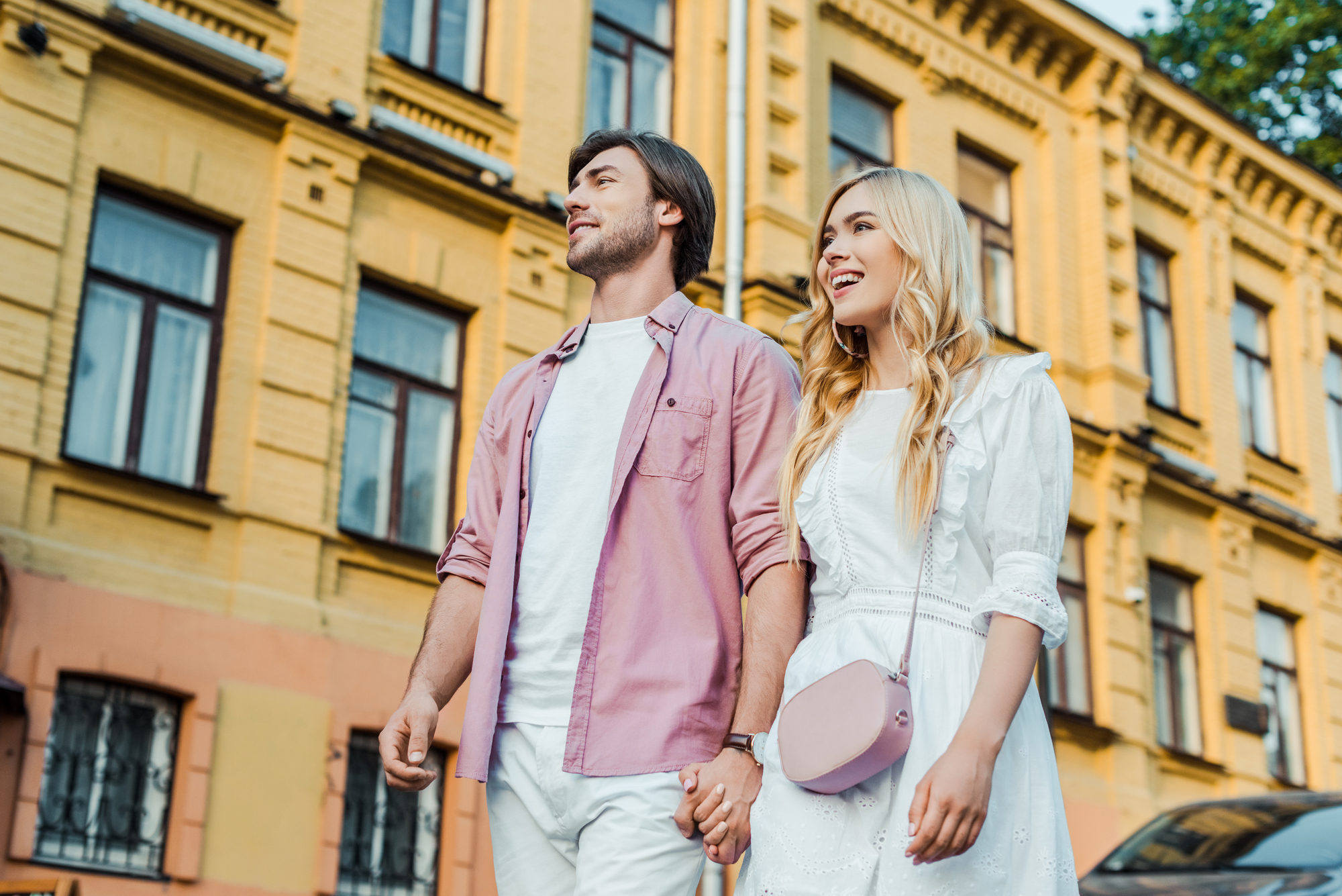 A smiling couple holds hands while walking down a street in front of yellow buildings. The man wears a light pink shirt and white pants, and the woman wears a white dress with a light pink purse. They appear happy and are enjoying the pleasant day.