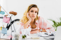 A woman with a frustrated expression sits at a cluttered desk with crumpled papers, a laptop, and a notebook. She rests her head on her hand, appearing tired or stressed. A bicycle, a mug, and some plants are visible in the background.