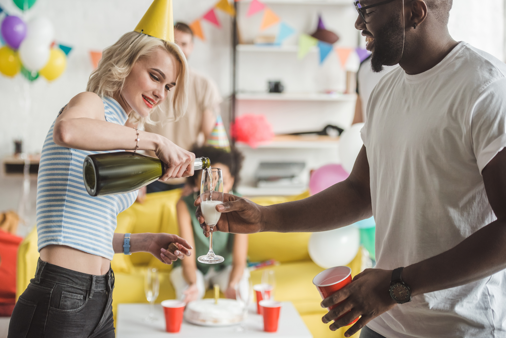 A woman in a birthday hat pours champagne into a man's glass at a festive party. They are standing in a colorful room with balloons, bunting, and a yellow couch in the background. Other guests are visible, enjoying the celebration.