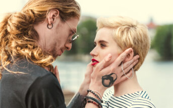 A couple gazes into each other's eyes with intense emotion. The man with long hair holds the woman's face tenderly. The woman has short blonde hair and bright red lipstick. They are standing outdoors, with blurred greenery and water in the background.