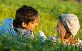 A young couple lies in a grassy field, facing each other and smiling warmly. The person on the left has short dark hair, while the person on the right wears a grey beanie and has long hair. The sunlight casts a soft, golden glow over them and the lush green grass.