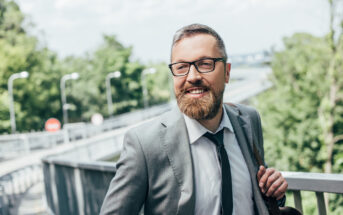 A bearded man with glasses wearing a gray suit and black tie stands smiling on a bridge in an outdoor setting with green foliage and a bright sky in the background.