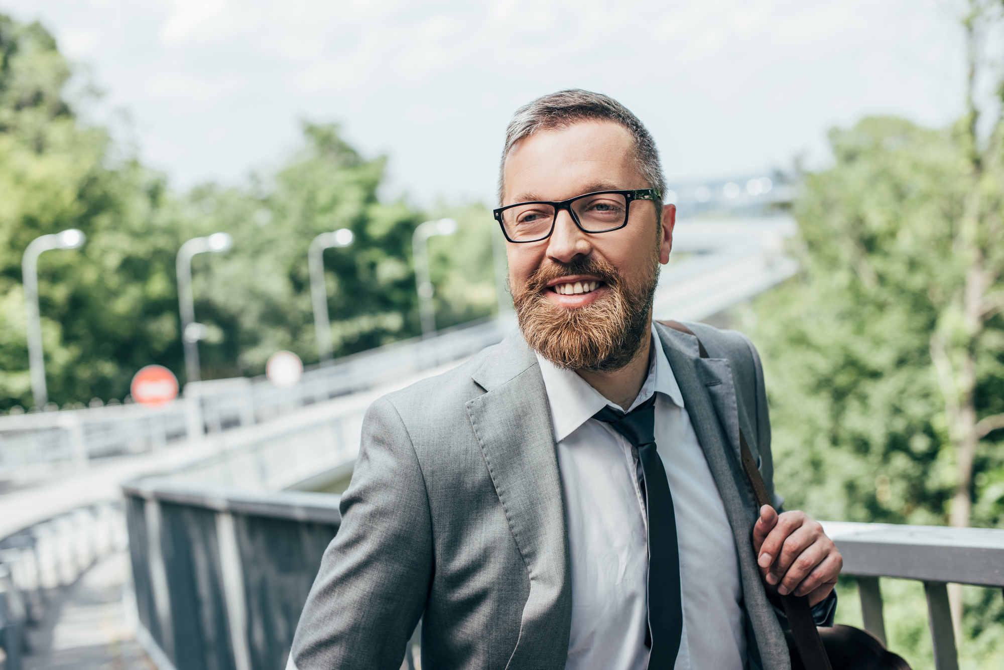 A bearded man with glasses wearing a gray suit and black tie stands smiling on a bridge in an outdoor setting with green foliage and a bright sky in the background.