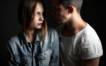 A young woman and man face each other closely against a dark background. The woman, wearing a denim jacket, looks to the side with a serious expression, while the man in a white t-shirt gazes intently at her. Their expressions suggest a tense or intense moment.