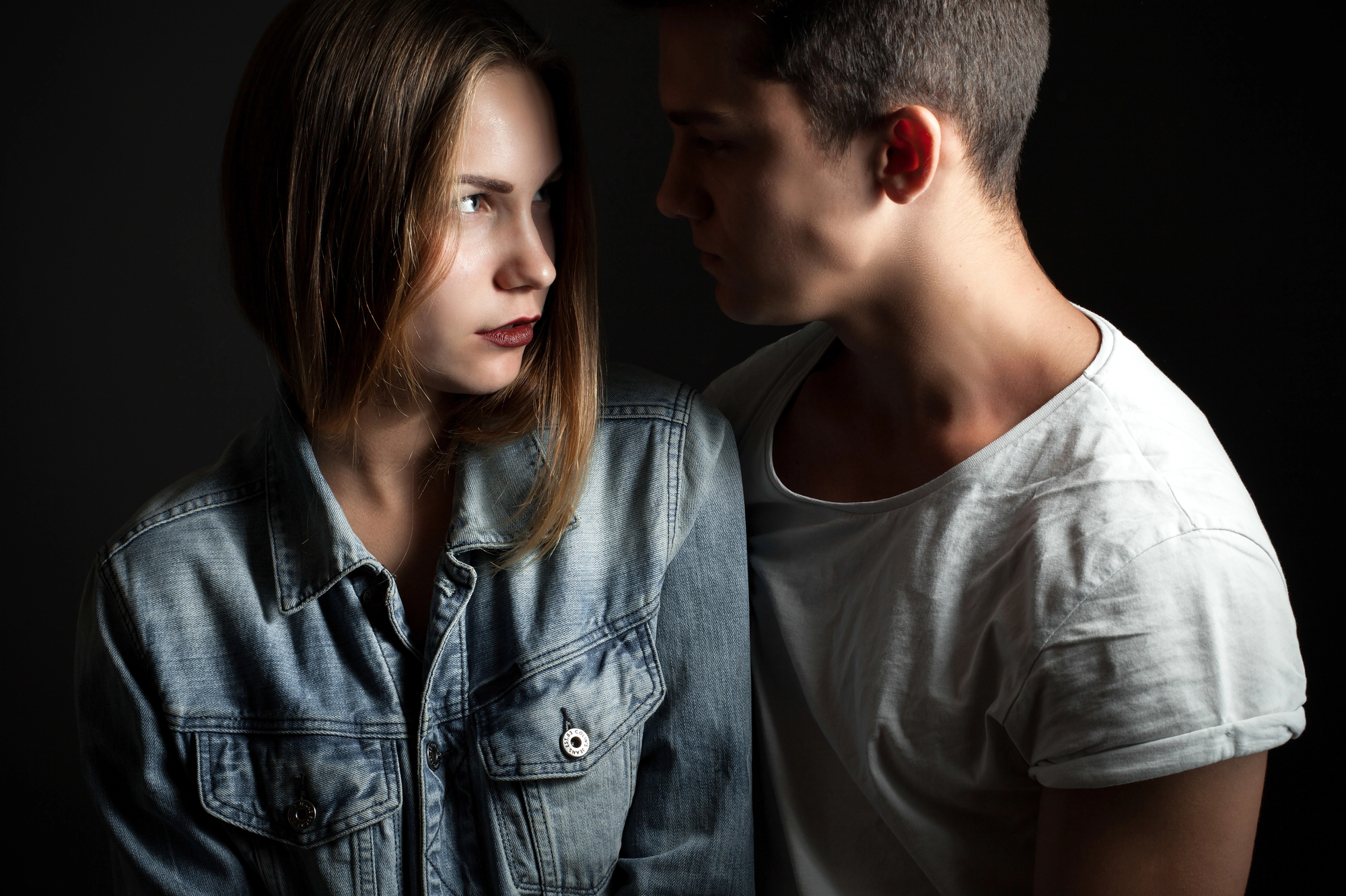 A young woman and man face each other closely against a dark background. The woman, wearing a denim jacket, looks to the side with a serious expression, while the man in a white t-shirt gazes intently at her. Their expressions suggest a tense or intense moment.