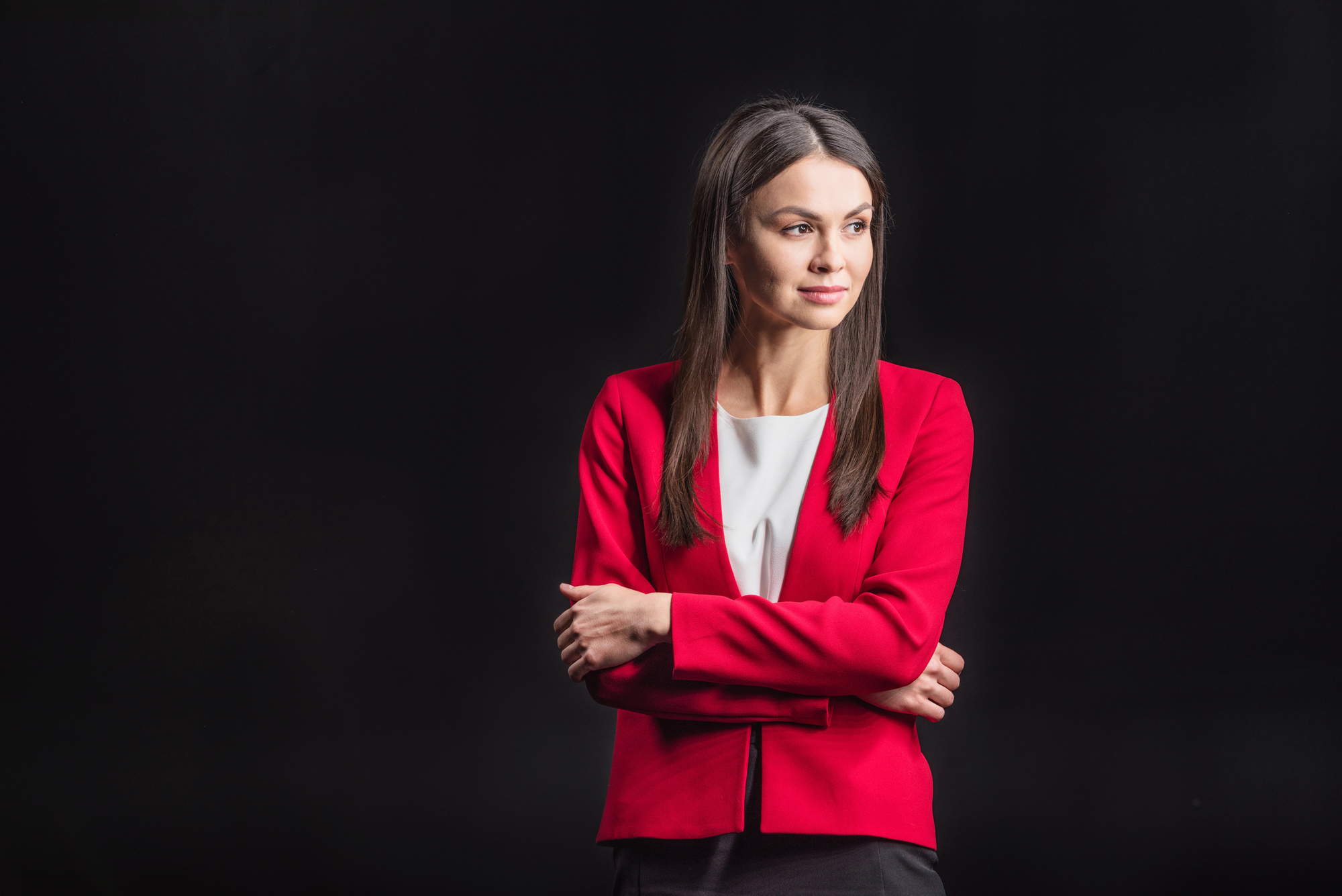 A woman with straight, dark hair is wearing a red blazer over a white top. She stands against a dark background with her arms crossed, looking slightly to her right with a thoughtful expression.