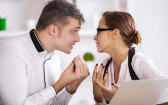 A man and a woman in a heated discussion at a desk in an office. The man is wearing a white shirt and gesturing with his hands, leaning towards the woman. The woman, also in a white shirt and wearing glasses, is responding with an intense expression. A laptop is open in front of them.